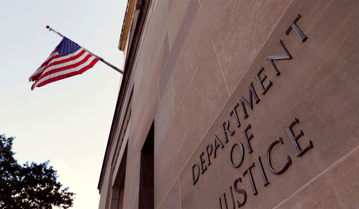 Photo of the outside of the US Department of Justice building. Logo on building offset. American flag above blows in the wind.