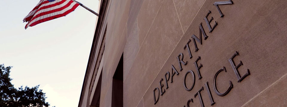 Photo of the outside of the US Department of Justice building. Logo on building offset. American flag above blows in the wind.