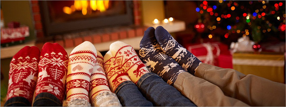 Four pairs of feet wearing festive holiday socks of different designs and colors rest on a coffee table in front of a roaring fireplace which sits just next to a beautiful Christmas Tree. Lights top to bottom. Gifts all around. Merry Christmas and Happy Holidays from Access Ready, Inc.