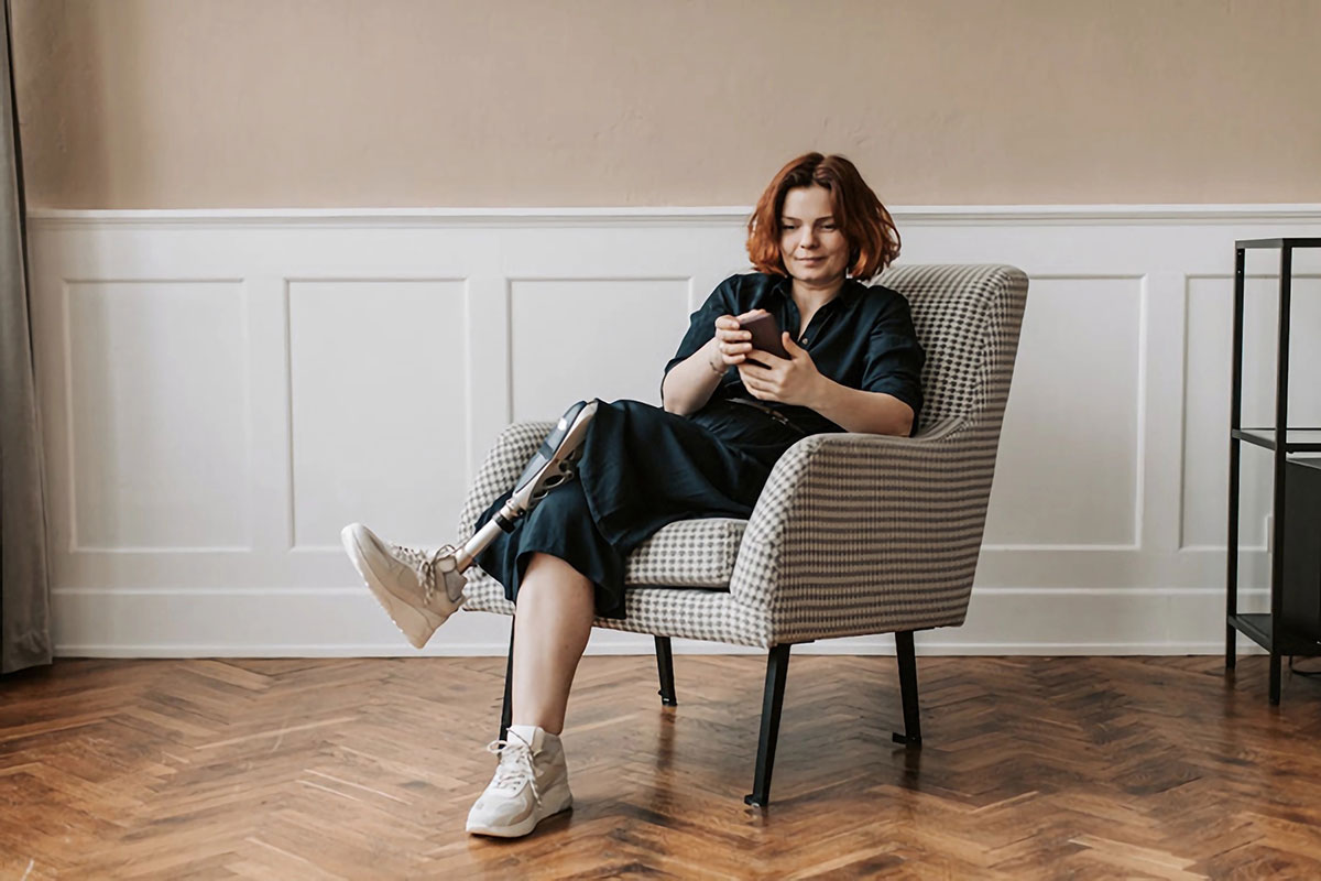 Planning Financially for the Future While Living with a Disability - Access Information News - A Mind Vault Solutions, Ltd. Publication A woman with a prosthetic leg is sitting comfortably in a patterned armchair in a well-lit room with light-colored walls and wooden flooring. She has shoulder-length auburn hair and is dressed in a dark outfit with white sneakers. She is smiling and looking at her smartphone, which she holds in both hands. The scene conveys a sense of relaxation and contentment.