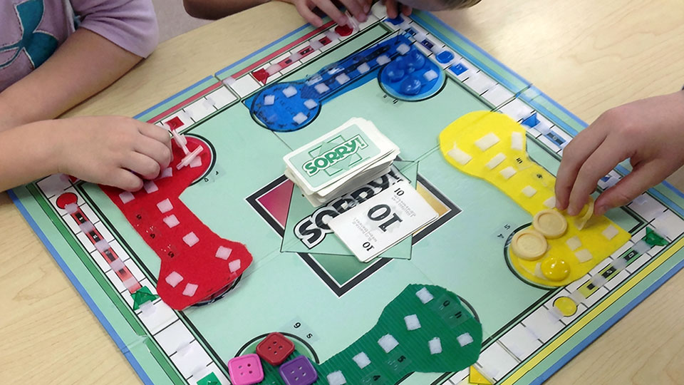 Children are playing a tactile version of the board game Sorry! on a light green game board with raised and textured features. The four player areas are colored red, blue, yellow, and green, each with velcro pieces and matching game pawns. The Sorry! card deck sits in the center, and the children are touching and moving the pieces around the board with their hands.