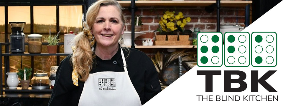 Debra Erickson, a woman with long blonde hair wearing a black chef's jacket and a white apron, stands smiling in a cozy kitchen with shelves of plants, jars, and cooking tools against a brick wall. The apron bears the logo of The Blind Kitchen, which is also displayed on the right side of the image, featuring the initials TBK in bold letters beneath three green braille-style icons and the text 'The Blind Kitchen'.
